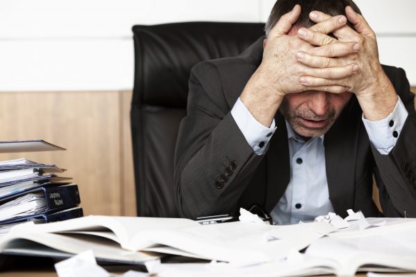 Worried businessman sitting at office desk full with books and papers being overloaded with work.